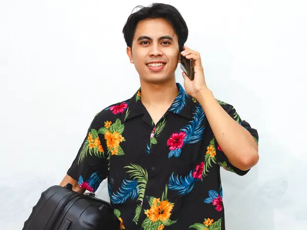 stock image Portrait of a smiling young Asian man in summer clothes holding a phone and suitcase, gesturing a call sign. Depicts a tourist ready for an overseas trip, isolated on white.