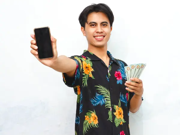 stock image A happy young Asian man in summer casual attire shows a blank mobile phone screen while holding cash, smiling against a clean white background.