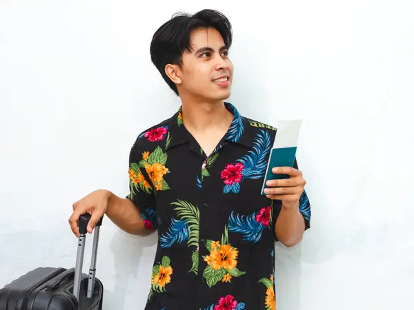 Stock image Cheerful young Asian man in summer casual attire holding a passport and suitcase, ready for a trip. Isolated on a white background, perfect for travel-themed content.