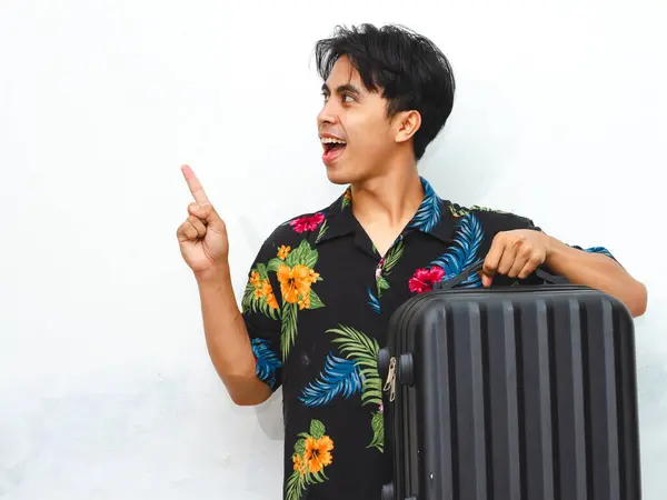 stock image Portrait of a young Asian man in casual summer clothes carrying a suitcase, pointing aside with a surprised expression. Ideal for travel, tourism, or vacation-related ads.