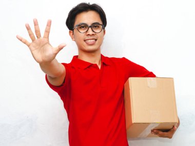 Kırmızı üniformalı, gülümseyen, kendine güveni tam bir Asyalı teslimatçının portresi. Elinde bir paket var ve beyaz arka planda beş parmağını gösteriyor. Teslimat veya kurye kavramları için ideal.