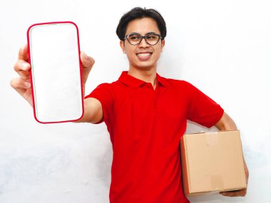 Paket taşıyan gülümseyen Asyalı bir teslimatçının portresi beyaz arka planda boş bir akıllı telefon ekranı gösteriyor. Teslimat kavramları için ideal..