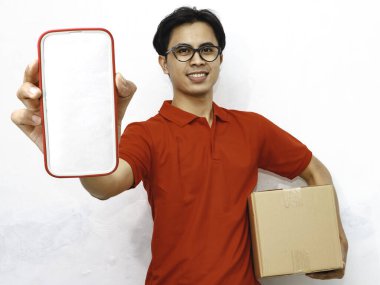Paket taşıyan gülümseyen Asyalı bir teslimatçının portresi beyaz arka planda boş bir akıllı telefon ekranı gösteriyor. Teslimat kavramları için ideal..