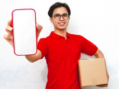 Portrait of a smiling friendly Asian delivery man holding a package while displaying a blank smartphone screen on an isolated white background, ideal for delivery concepts. clipart