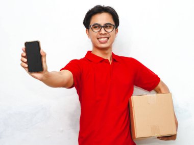 Paket taşıyan gülümseyen Asyalı bir teslimatçının portresi beyaz arka planda boş bir akıllı telefon ekranı gösteriyor. Teslimat kavramları için ideal..