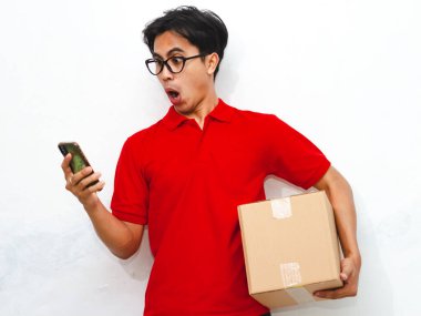 Paket taşıyan ve akıllı telefon kullanan Asyalı bir kuryenin portresi beyaz arka planda komik bir şok ifadesi gösteriyor. Teslimat veya kurye kavramları için ideal.