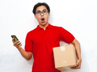 Paket taşıyan ve akıllı telefon kullanan Asyalı bir kuryenin portresi beyaz arka planda komik bir şok ifadesi gösteriyor. Teslimat veya kurye kavramları için ideal.