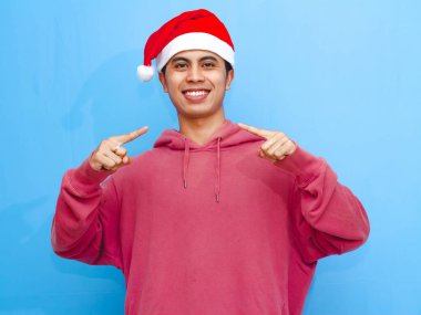 Happy young Asian man in a red hoodie and Santa hat, confidently pointing at himself, perfect for Christmas and New Year advertising themes