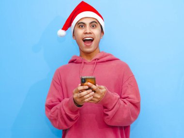 Kırmızı kapüşonlu ve Noel Baba şapkalı neşeli bir Asyalı adamın portresi. Elinde tuttuğu ve şaşırmış bir ifadeyle akıllı telefonu kullanan. Noel ve Yeni Yıl reklam kavramları için ideal.