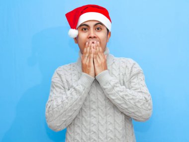 Süveter giyen genç bir Asyalı adam portresi ve Noel Baba şapkası, elleriyle ağzını kapatıyor, şaşırmış bir ifade sergiliyor. Sade mavi bir arkaplanda izole edilmiş.