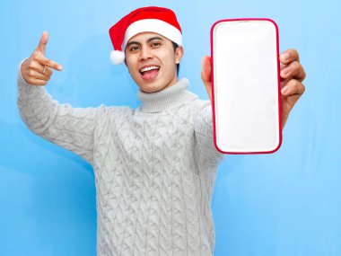 Süveter ve Noel Baba şapkalı mutlu Asyalı genç bir adamın portresi, boş bir akıllı telefon ekranını heyecanla gösteriyor. Sade mavi bir arka planda izole edilmiş, tatil reklamları için mükemmel..