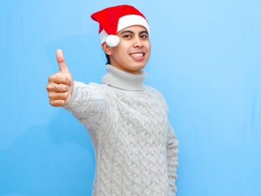 Portrait of a happy young Asian man wearing a sweater and Santa hat, giving a thumbs up on a plain blue background. Celebrating New Year and the holiday season in a joyful mood.