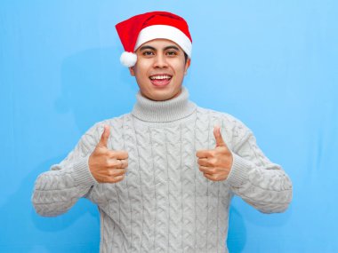 Portrait of a happy young Asian man wearing a sweater and Santa hat, giving a thumbs up on a plain blue background. Celebrating New Year and the holiday season in a joyful mood.