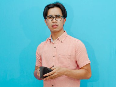 Pembe gömlekli Asyalı bir adam boş bir cüzdan tutuyor ve izole edilmiş mavi arka planda inanılmaz bir yüz ifadesi gösteriyor. Mali sıkıntı, borç veya kayıp gibi kavramlar için ideal.