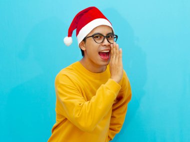 Asyalı bir adam Noel şapkası takıyor, neşeli bir ifadeyle bir şeyler fısıldıyor. Bayram reklamları, tatil promosyonları ve neşeli mevsimsel içerik için mükemmel..