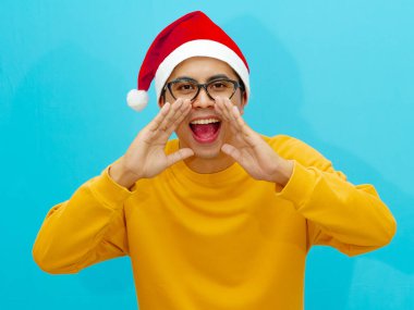 Genç Asyalı bir adam Noel şapkası takıp elleri ağzının kenarında bağırıyor, satışlar ve indirimler hakkındaki haberleri heyecanla paylaşıyor. Tatil temalı reklamlar ve promosyonlar için ideal..