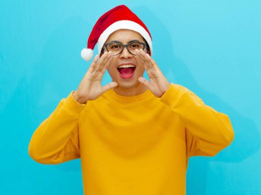 Genç Asyalı bir adam Noel şapkası takıp elleri ağzının kenarında bağırıyor, satışlar ve indirimler hakkındaki haberleri heyecanla paylaşıyor. Tatil temalı reklamlar ve promosyonlar için ideal..