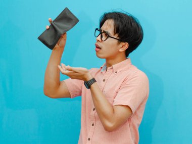 Pembe gömlekli Asyalı bir adam boş bir cüzdan tutuyor ve izole edilmiş mavi arka planda inanılmaz bir yüz ifadesi gösteriyor. Mali sıkıntı, borç veya kayıp gibi kavramlar için ideal.