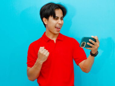 Kırmızı polo tişörtlü heyecanlı bir Asyalı adamın portresi zaferi kutlarken mavi arka planda izole edilmiş, reklam ya da promosyon için mükemmel bir oyun akışı izliyor..