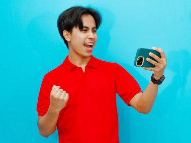 Kırmızı polo tişörtlü heyecanlı bir Asyalı oyuncunun portresi gezici bir oyun oynarken zaferi kutluyor. Mavi bir arkaplanda izole edilmiş, coşku ve başarı gösteriyor..