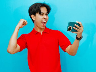 Kırmızı polo tişörtlü heyecanlı bir Asyalı oyuncunun portresi gezici bir oyun oynarken zaferi kutluyor. Mavi bir arkaplanda izole edilmiş, coşku ve başarı gösteriyor..