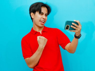 Kırmızı polo tişörtlü heyecanlı Asyalı adamın portresi yumruğunu kaldırıyor ve maçı cep telefonuyla kazanıyor..