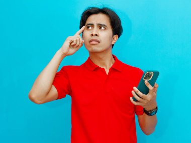 Kırmızı polo tişörtlü kafası karışmış Asyalı bir adam elinde sinirli ve stresli bir ifadeyle cep telefonu tutuyor. Mavi bir arkaplanda izole edilmiş.