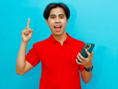 Kırmızı polo tişörtlü heyecanlı bir Asyalı adam, elinde bir telefon tutarken, bir fikir ediniyormuş gibi, mavi arka planda izole edilmiş. Reklam, yaratıcılık ve yenilik kavramları için mükemmel..
