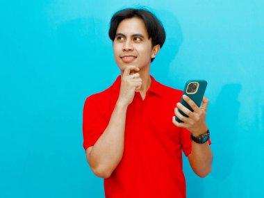 Kırmızı polo tişörtlü gülümseyen Asyalı bir adamın portresi cep telefonu tutarken mavi bir arkaplanda, düşünürken bir yüz ifadesi gösteriyor. Reklam için mükemmel..