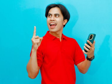 Kırmızı polo tişört giyen Asyalı bir adamın portresi, elinde cep telefonu tutarken mutlu ve heyecanlı bir ifadeyle, mavi arka planda izole edilmiş, reklam için mükemmel..