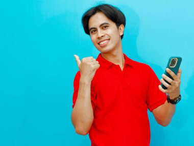 Kırmızı polo tişörtlü gülümseyen Asyalı bir adamın portresi, bir cep telefonu tutuyor ve başparmağıyla işaret ediyor, izole edilmiş mavi bir arka planda, reklam veya promosyon için ideal..