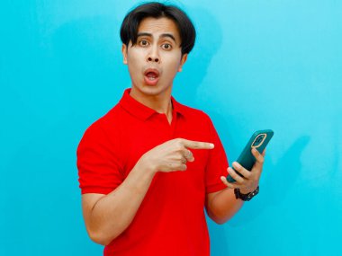 Kırmızı polo tişörtlü Asyalı bir adamın portresi mavi izole bir arkaplanda şok edici bir ifadeyle cep telefonu tutuyor. Samimi bir anda çekilen komik bir tepki..