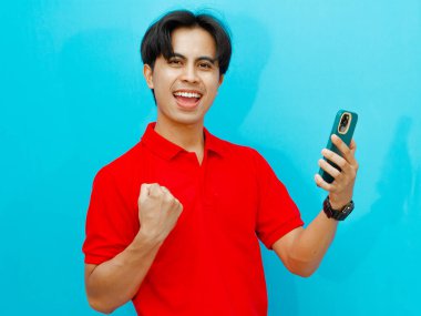 Kırmızı polo tişörtlü heyecanlı bir Asyalı adamın portresi başarıyı kutlarken bir yandan da cep telefonunu izole edilmiş mavi arka planda tutuyor..