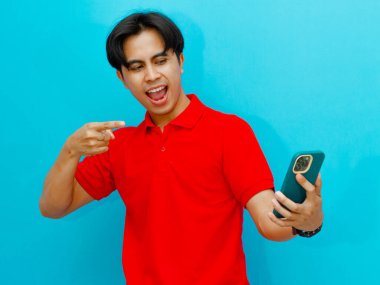Kırmızı polo tişört giyen heyecanlı bir Asyalı adamın portresi, cep telefonuna hevesli bir ifadeyle bakıyor, mavi arka planda izole edilmiş. Teknoloji ve reklam temaları için mükemmel.