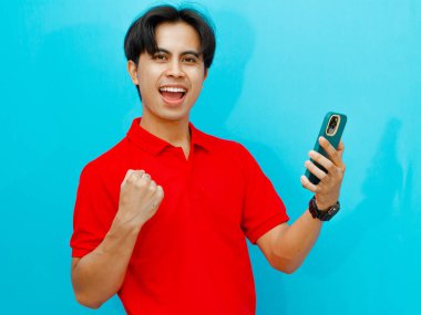 Kırmızı polo tişörtlü heyecanlı bir Asyalı adamın portresi başarıyı kutlarken bir yandan da cep telefonunu izole edilmiş mavi arka planda tutuyor..