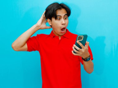 Kırmızı polo tişörtlü Asyalı bir adamın portresi mavi izole bir arkaplanda şok edici bir ifadeyle cep telefonu tutuyor. Samimi bir anda çekilen komik bir tepki..