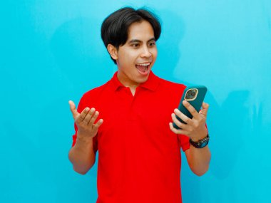 Kırmızı polo tişörtlü Asyalı bir adamın portresi, cep telefonunu izole edilmiş mavi arka planla karşılaştırırken şaşırmış görünüyor..