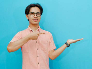 Pembe gömlekli arkadaş canlısı Asyalı bir adamın portresi. Mavi arka planda izole edilmiş bir ürün ya da reklamı sunmak ya da sergilemek için avucunun içini işaret ediyor ve gülümsüyor..