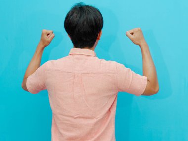 Rear view of an Asian man celebrating victory, raising his hand, and clenching his fist, showing excitement and success. Isolated on blue background, ideal for advertising and promotions. clipart