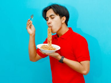 Kırmızı polo tişörtü giyen Asyalı bir adamın portresi, elinde bir tabak kızarmış şehriye, mavi arka planda mutlu bir ifadeyle yemeğinin tadını çıkarıyor..