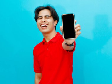Kırmızı polo tişörtlü Asyalı bir adam elinde boş ekranlı bir cep telefonu tutuyor ve izole edilmiş mavi arka planda gülüyor. Reklamcılık veya teknolojiyle ilgili kavramlar için mükemmel..