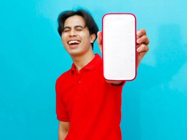 Kırmızı polo tişörtlü Asyalı bir adam elinde boş ekranlı bir cep telefonu tutuyor ve izole edilmiş mavi arka planda gülüyor. Reklamcılık veya teknolojiyle ilgili kavramlar için mükemmel..