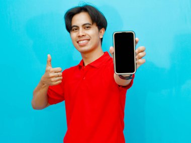 Kırmızı polo tişörtlü Asyalı bir adamın portresi boş bir cep telefonu ekranı tutuyor ve mavi arka planda izole edilmiş gülümseyen bir yüzle baş parmak kaldırıyor..