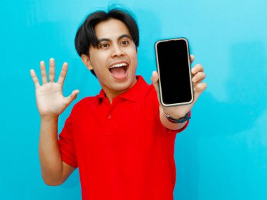 Kırmızı polo tişört giyen Asyalı bir adamın portresi, mavi arka planda izole edilmiş şok bir ifadeyle boş bir cep telefonu ekranını gösteriyor..