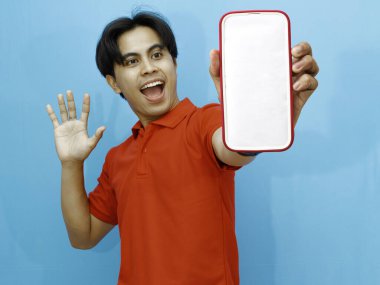 Kırmızı polo tişört giyen Asyalı bir adamın portresi, mavi arka planda izole edilmiş şok bir ifadeyle boş bir cep telefonu ekranını gösteriyor..