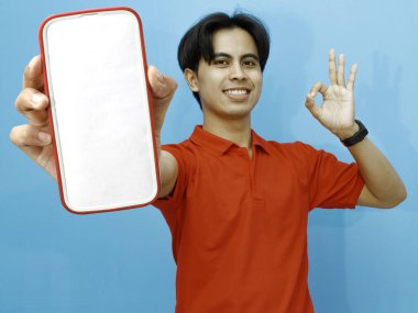 Kırmızı polo tişörtlü Asyalı bir adamın portresi boş bir cep telefonu ekranı sergiliyor, mavi arka plana karşı heyecanlı bir ifadeyle işaret parmağı veriyor..