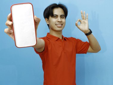 Kırmızı polo tişörtlü Asyalı bir adamın portresi boş bir cep telefonu ekranı sergiliyor, mavi arka plana karşı heyecanlı bir ifadeyle işaret parmağı veriyor..