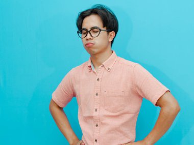 Sad adult Asian businessman in pink shirt standing with a stressed and despairing expression, isolated on a blue background. Ideal for concepts of failure, burnout, and workplace stress. clipart