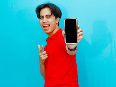 Kırmızı polo tişörtlü Asyalı bir adamın portresi mavi arka planda izole edilmiş mutlu bir ifadeyi gösteren boş bir cep telefonu ekranını işaret ediyor..