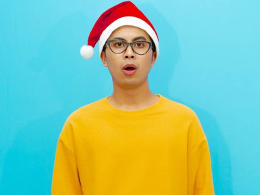 Asian man wearing a Christmas hat shows a funny shocked face expression while looking at the camera. clipart